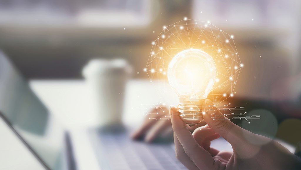 Onde encontrar ideias de conte&uacute;dos para Instagram? : m&atilde;o segurando uma l&acirc;mpada com luzes e um notebook de fundo