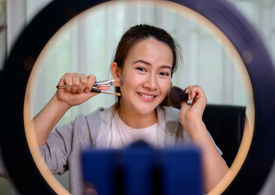 micro influencer asiatica femininacom pinceis encostando no rosto de frente uma luminária de maquiagem
