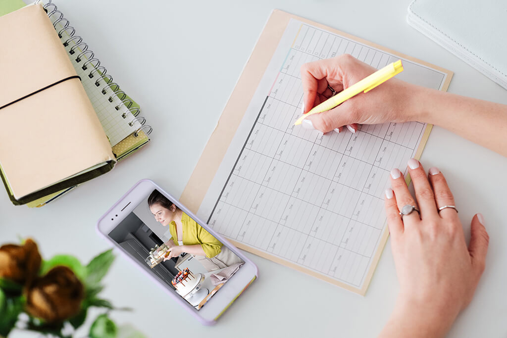 Como ser um influenciador digital? : Sob uma mesa um calendario com m&atilde;os a preench&ecirc;-lo, um celular, uma agenda e flores