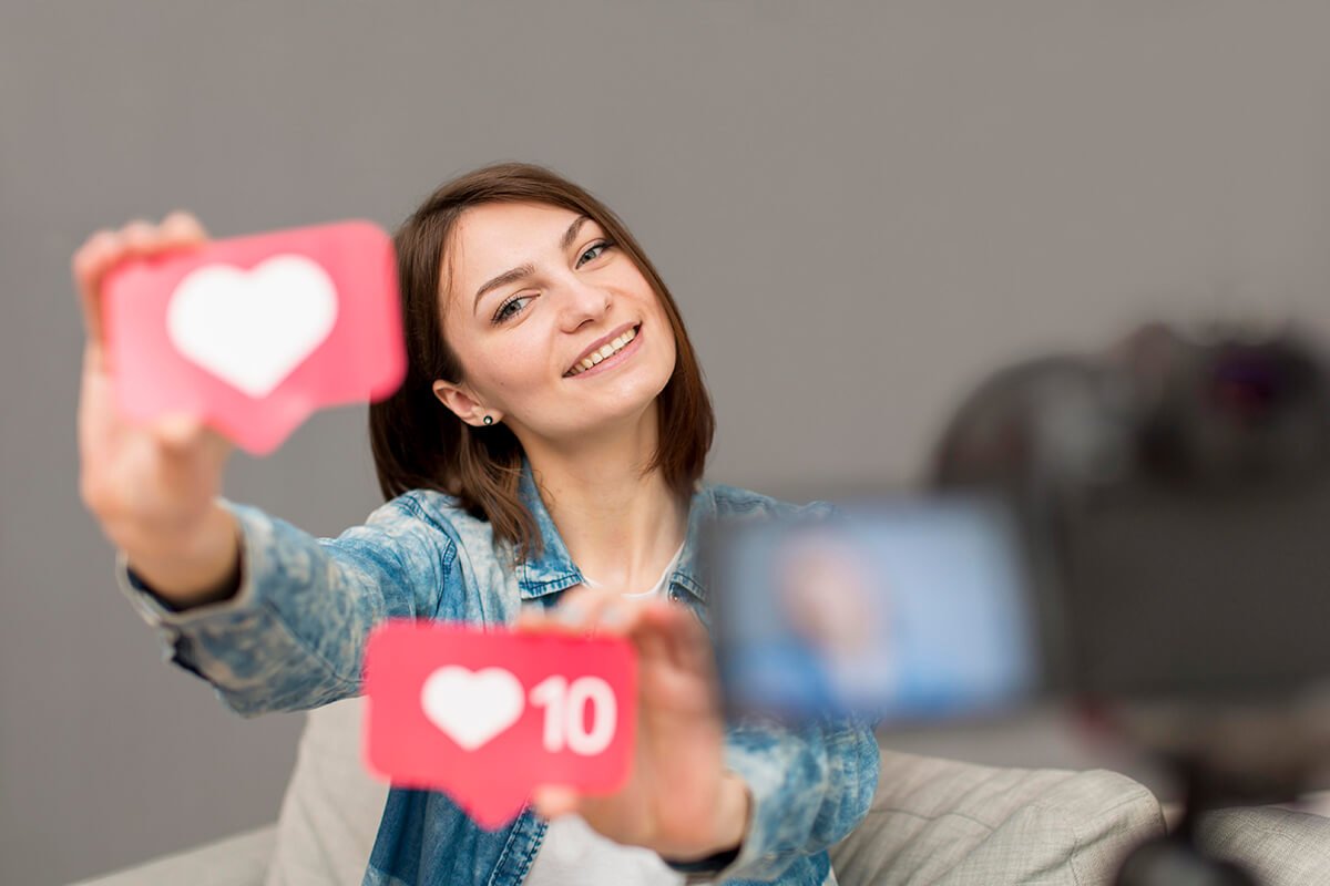 Imagem de uma influenciadora de frente &agrave;s c&acirc;meras, mostrando placas com curtidas das redes sociais, exemplificando o conceito da profiss&atilde;o que consiste em compartilhar conte&uacute;do, gerar opini&otilde;es e influenciar decis&otilde;es de compra. Fonte: @Freepik