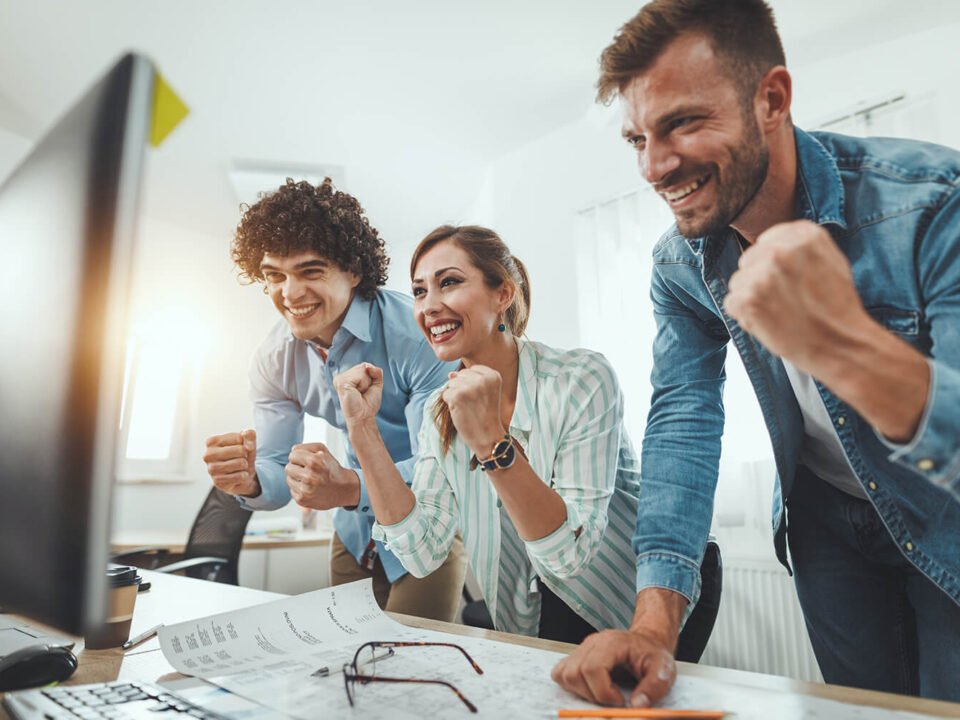 Capa eng.interno 3 funcionários comemorando vitória em frente uma mesa com copmputador e papéis