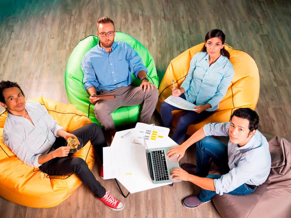 capa benef. gamif. 4 pessoas trabalhando e sentadas em pufs em volta de uma mesa com um computdor